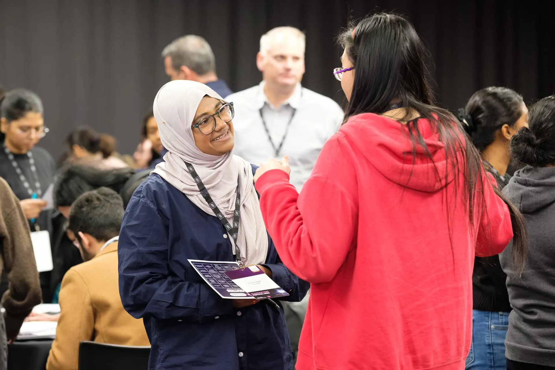 A woman in a hijab smiles while talking with another attendee, a printed handout in her hand, in a room full of people meeting each other.