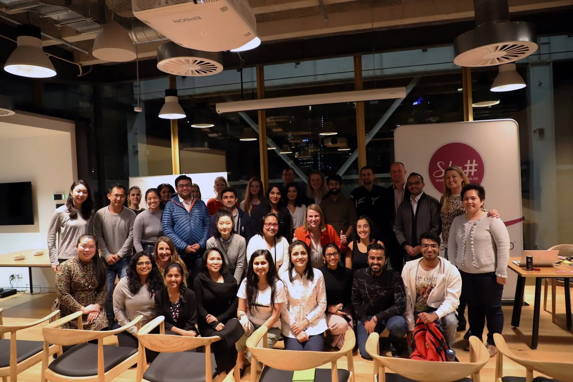 A group of She Sharp members gathered together, seated and standing, in a warmly lit lounge at an Amazon Web Services event.