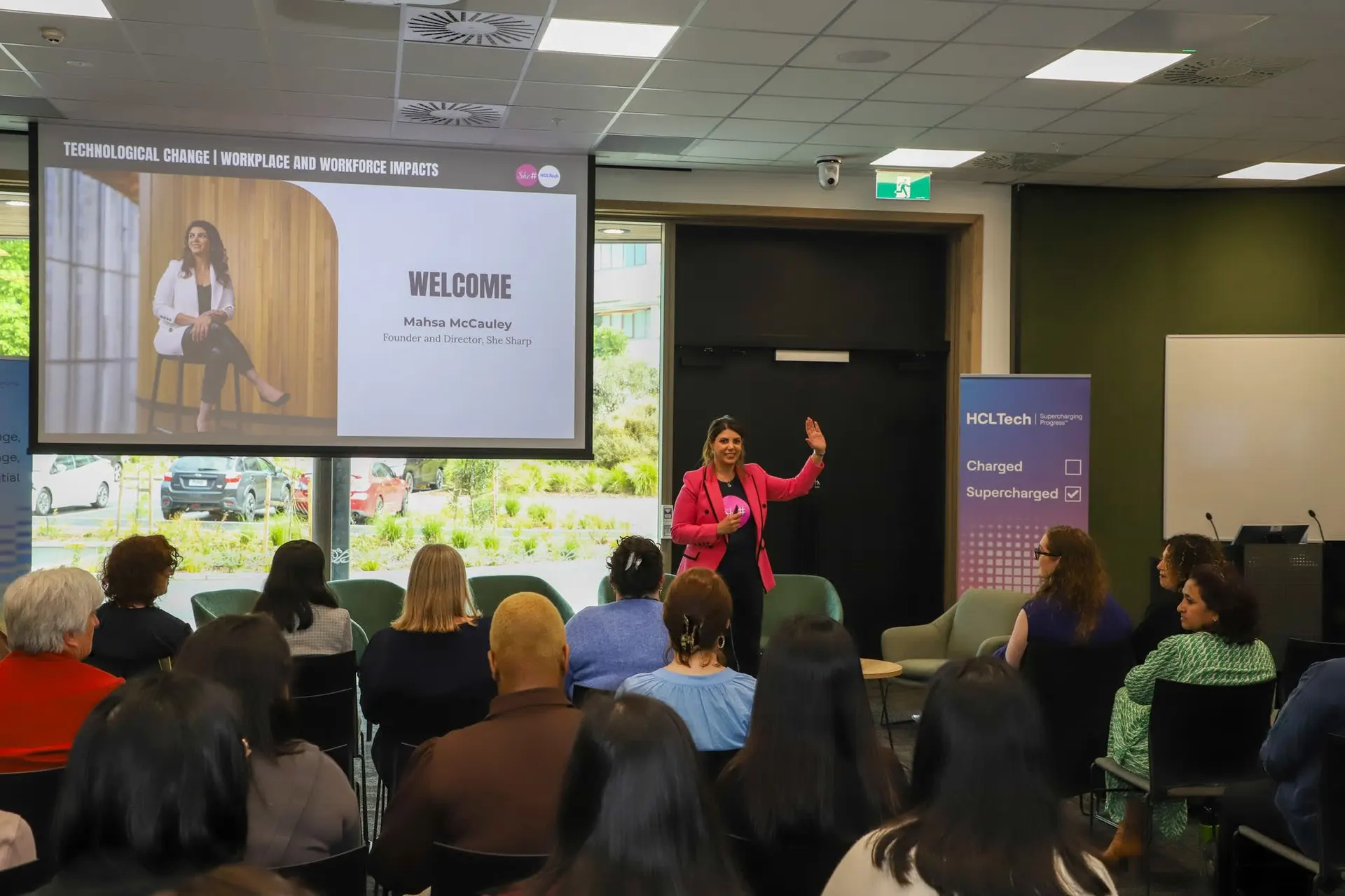 A presenter welcoming a seated audience at the start of a She Sharp event.