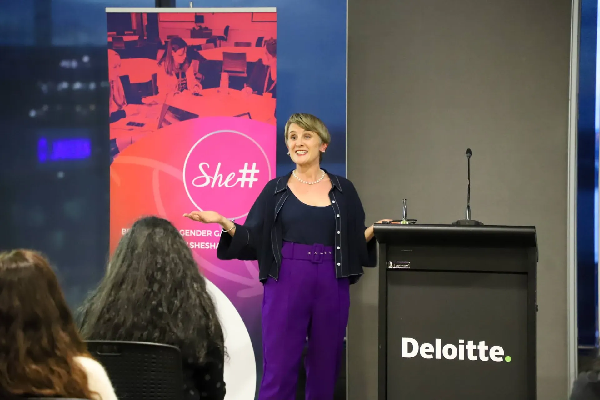 A She Sharp speaker presenting from a podium on a darkened stage.