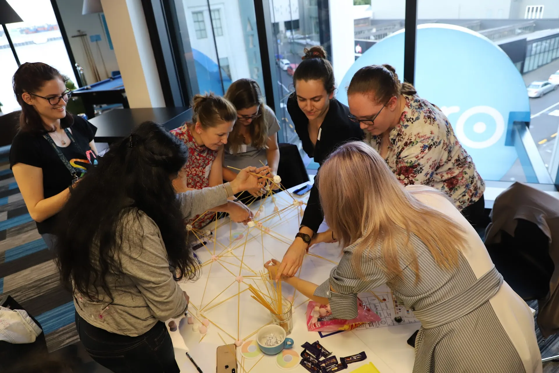 Participants leaning in together to build a spaghetti-and-tape tower during a hands-on She Sharp workshop challenge.