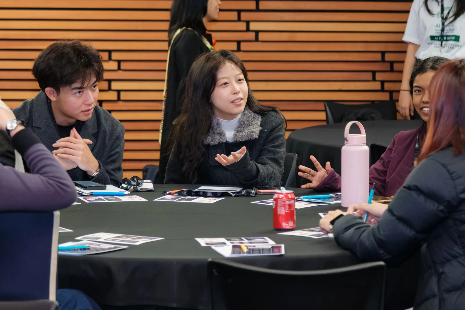 A participant speaks with an open hand across a round table while her teammates listen, printed prompt cards spread on the black tablecloth.