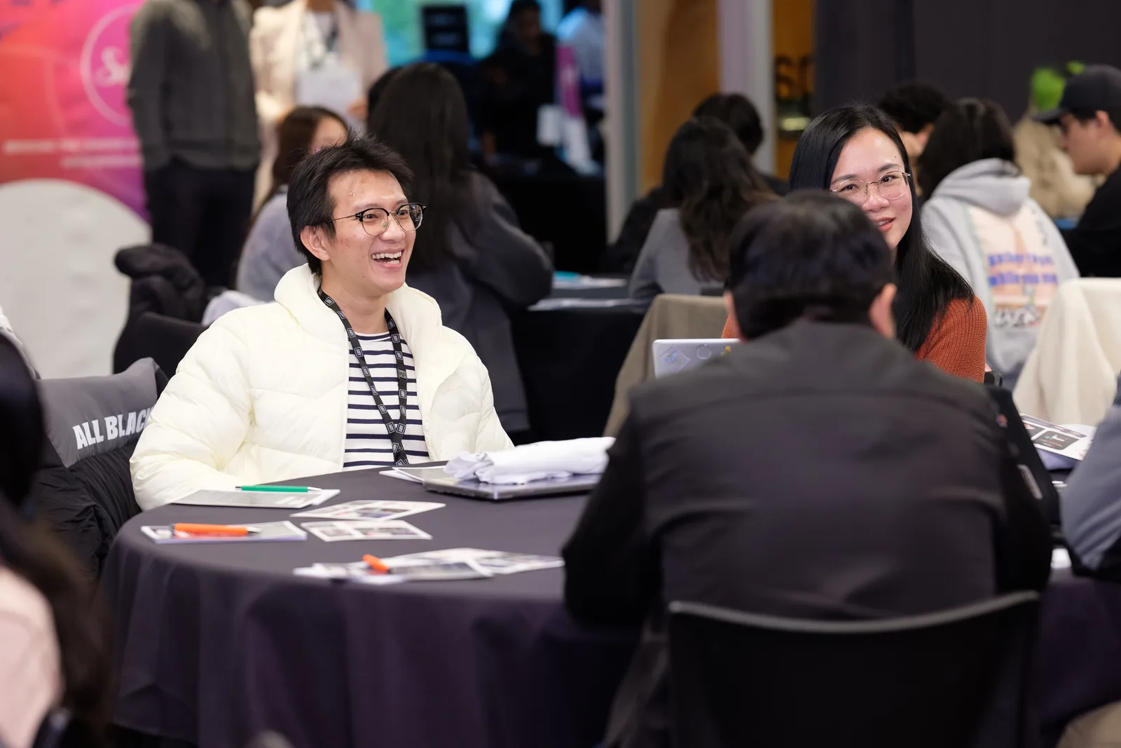 A participant in a cream puffer jacket laughs broadly across her table in a crowded working hall.