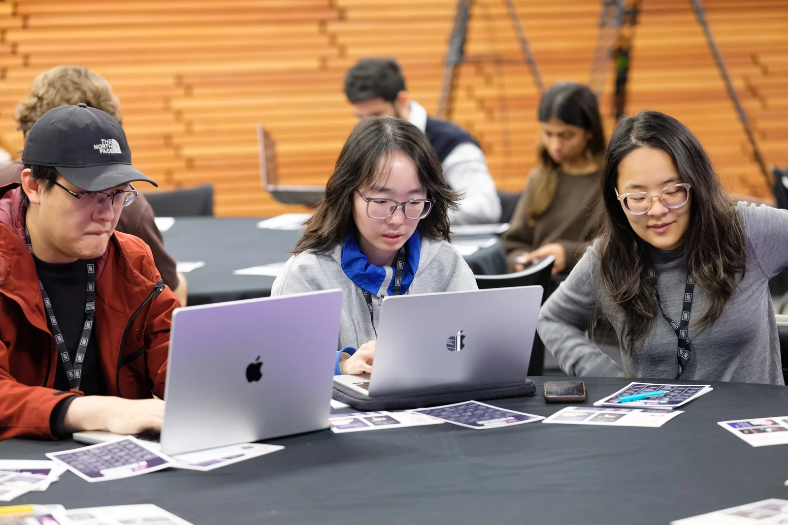 Three participants sit shoulder to shoulder over open laptops, concentrating, against a warm timber slat wall.