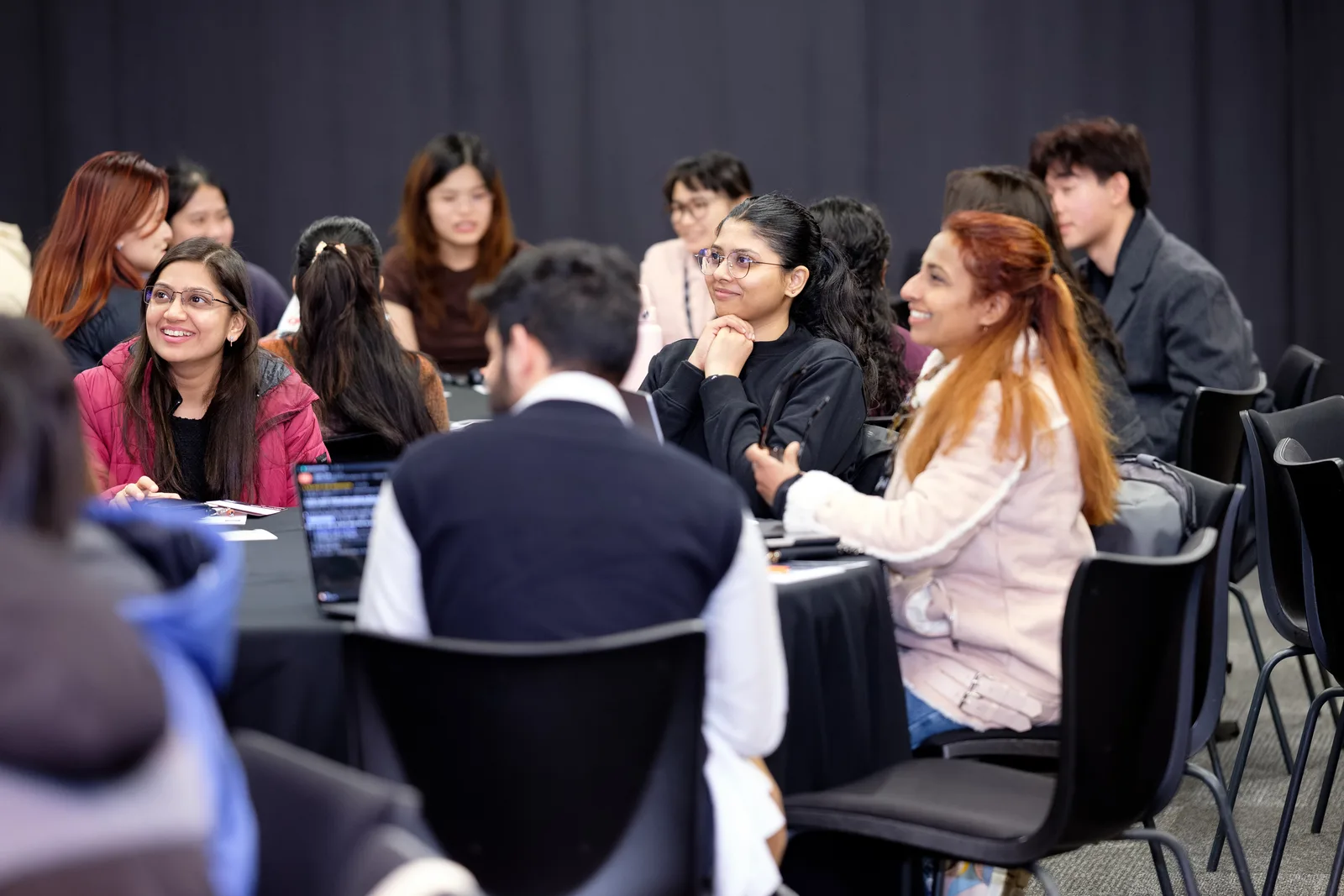 A round table of participants, several of them smiling, turn towards a speaker in a room hung with dark curtains.