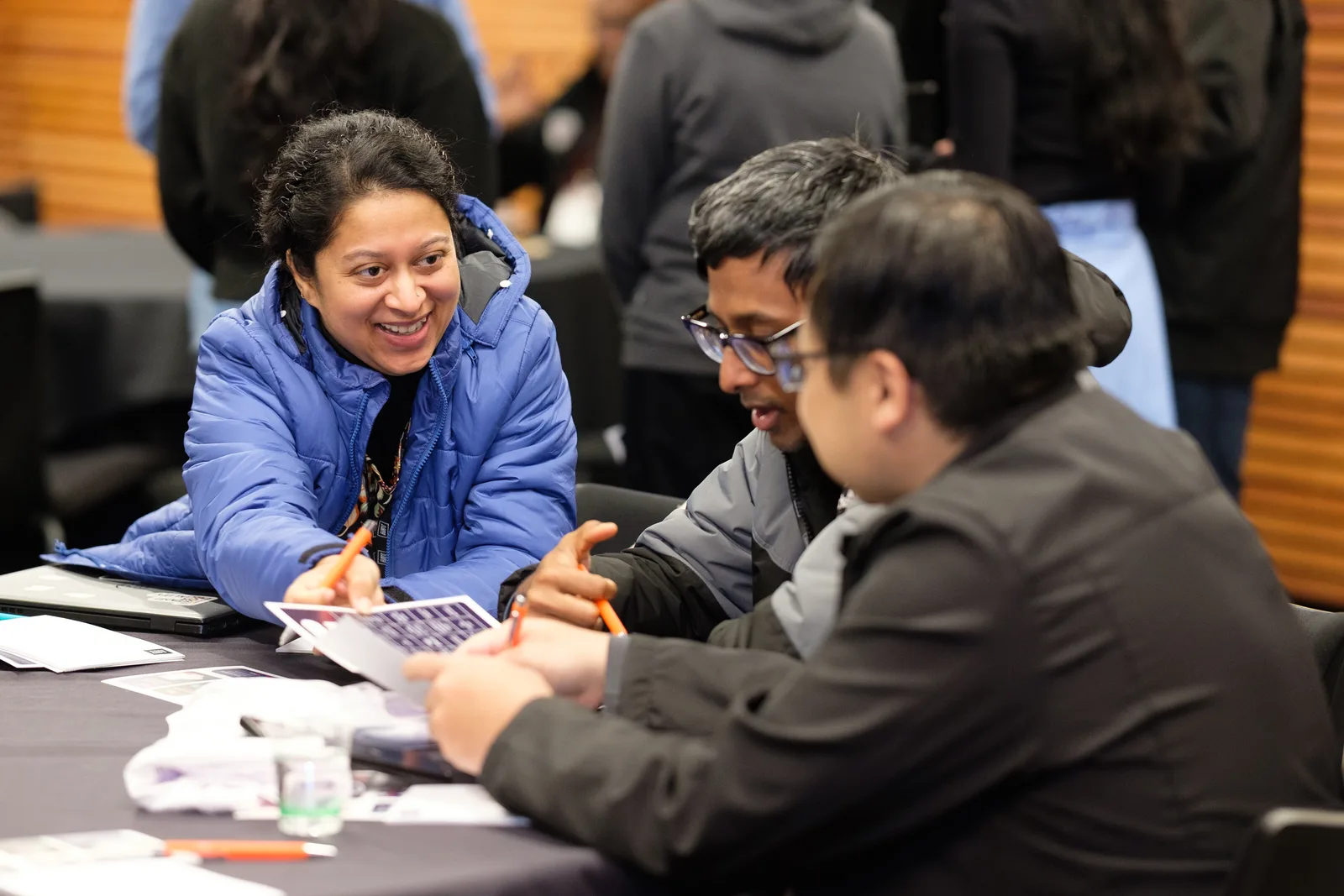 A woman in a blue puffer jacket laughs mid-conversation with two teammates working over notes at their table.