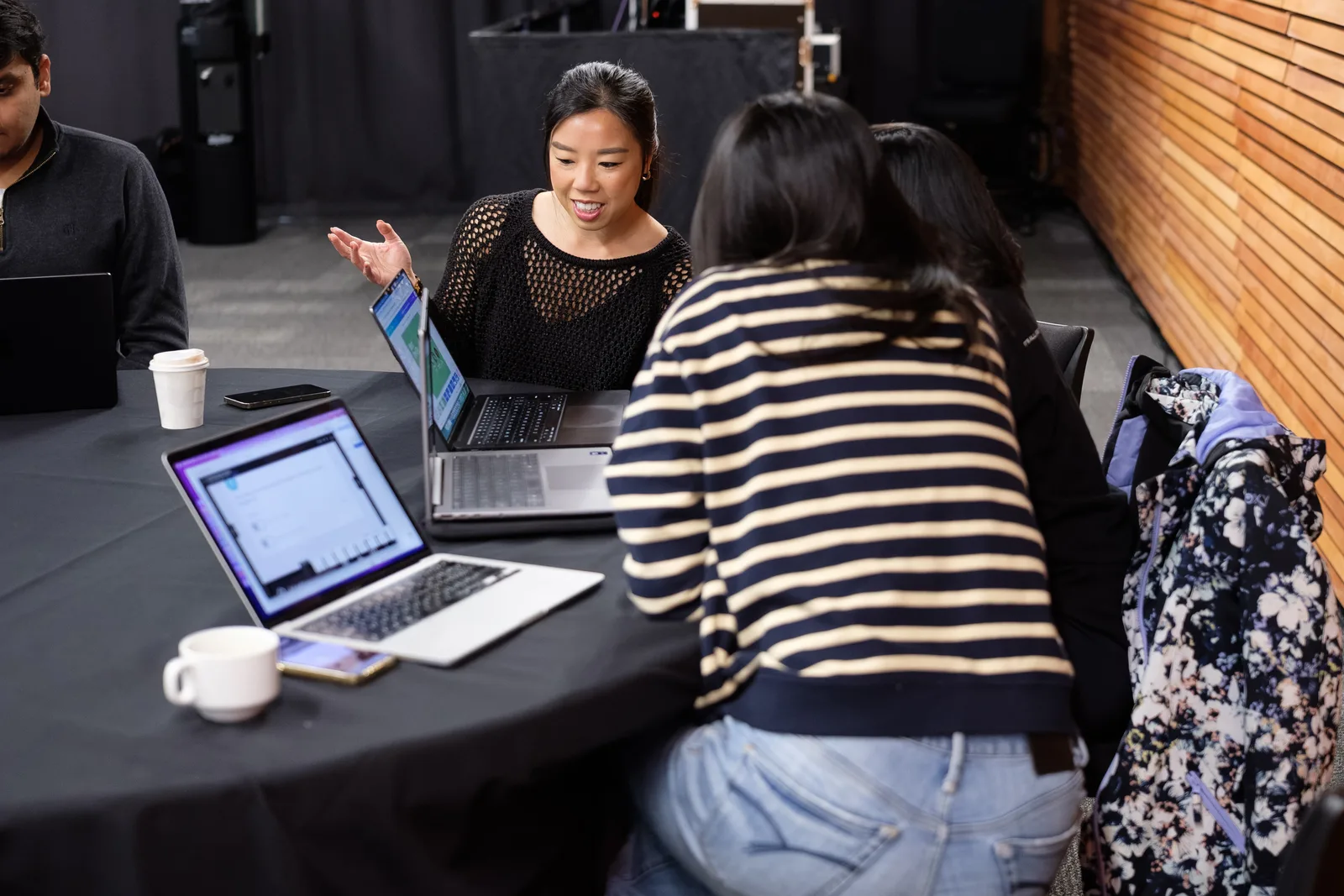 A woman gestures with an open hand as she explains something across a table of open laptops to two listening teammates.