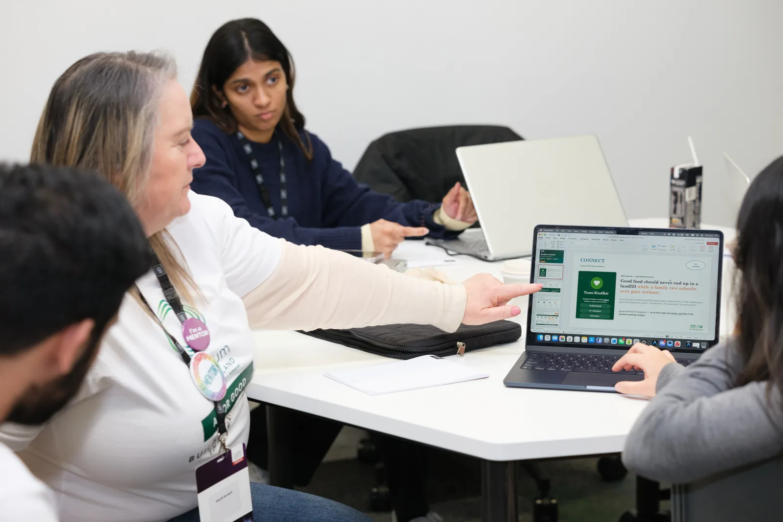 A mentor wearing an I'm a mentor badge reaches across the table to point at a teammate's laptop screen showing a team's pitch slide.