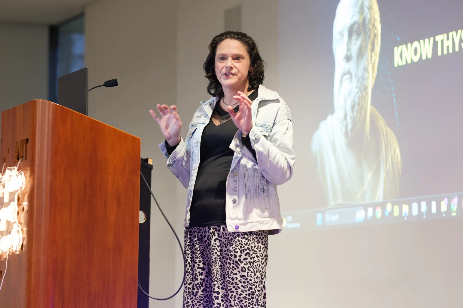 A keynote speaker raises both hands mid-sentence at a wooden AUT lectern, a classical marble bust and the words KNOW THYSELF projected behind her.