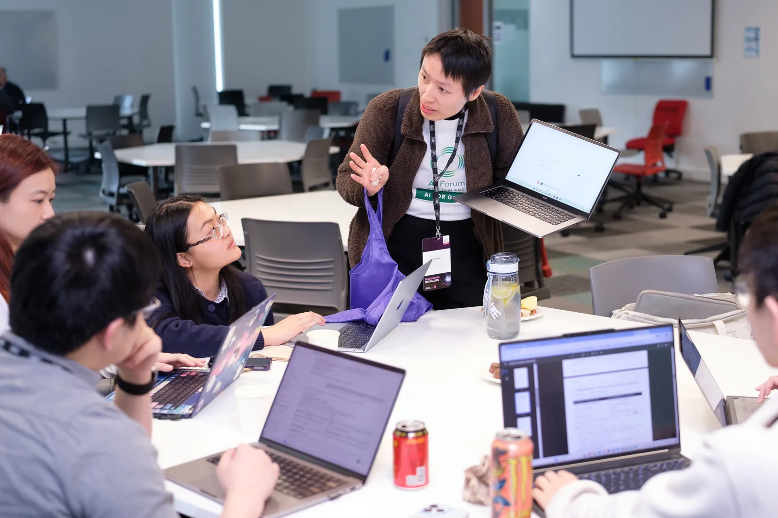 A mentor stands beside a table with a laptop in hand, talking a seated team through their idea