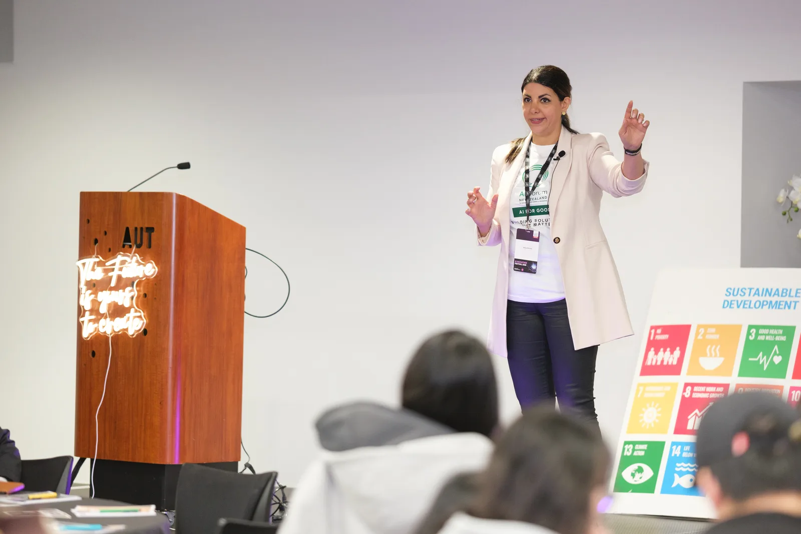 A host throws both arms wide beside an AUT lectern lit by a neon sign reading The Future is yours to create, a Sustainable Development Goals board to one side.