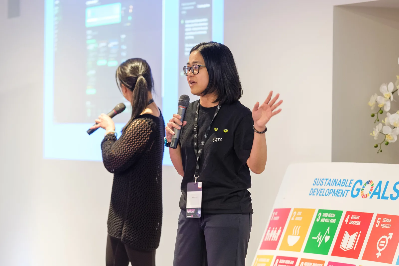 Two team members present on stage holding microphones, one gesturing mid-sentence, with a projected slide behind them and a Sustainable Development Goals board at their side.