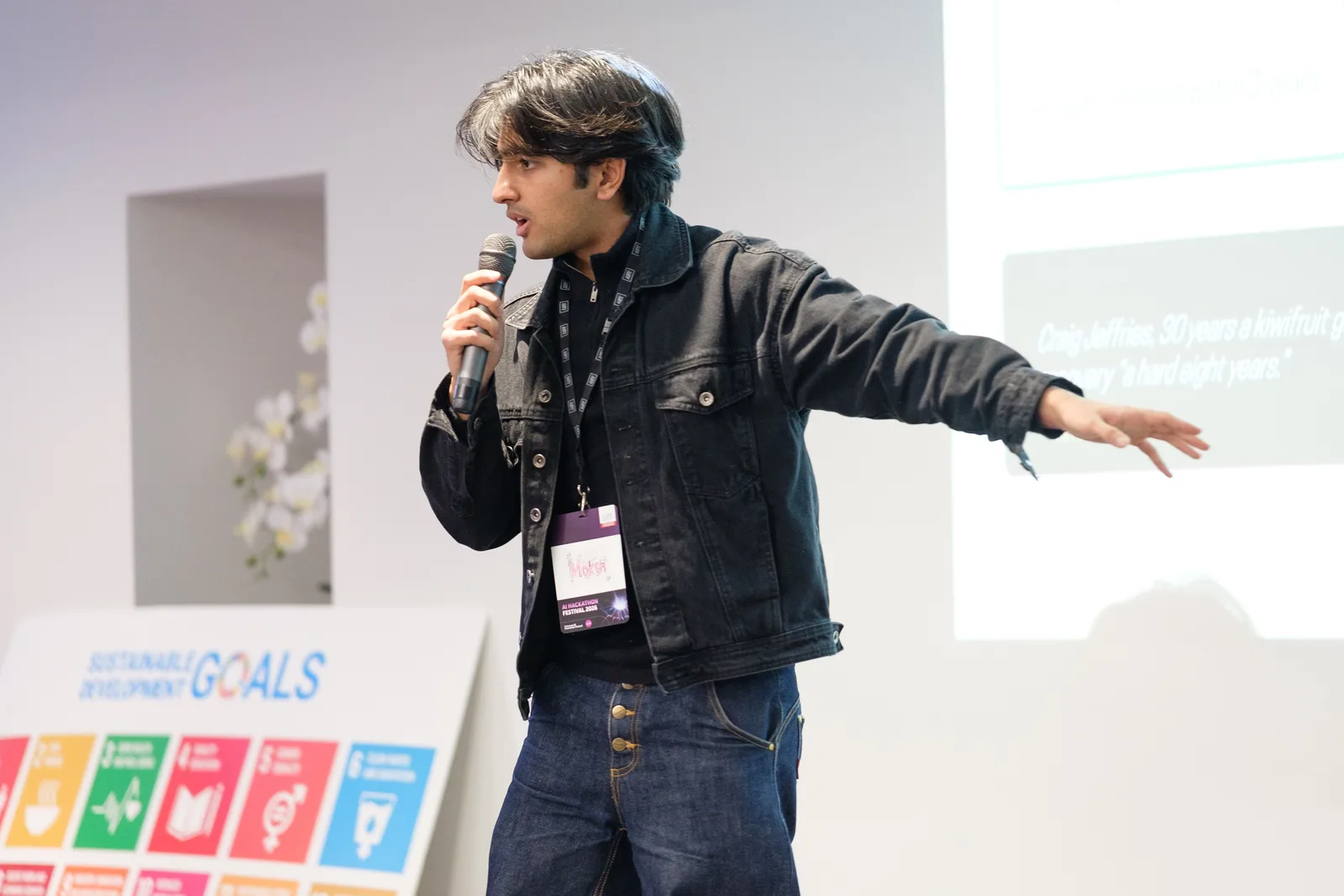 A presenter flings one arm wide mid-sentence with the microphone raised, the Sustainable Development Goals board standing at the edge of the stage behind.