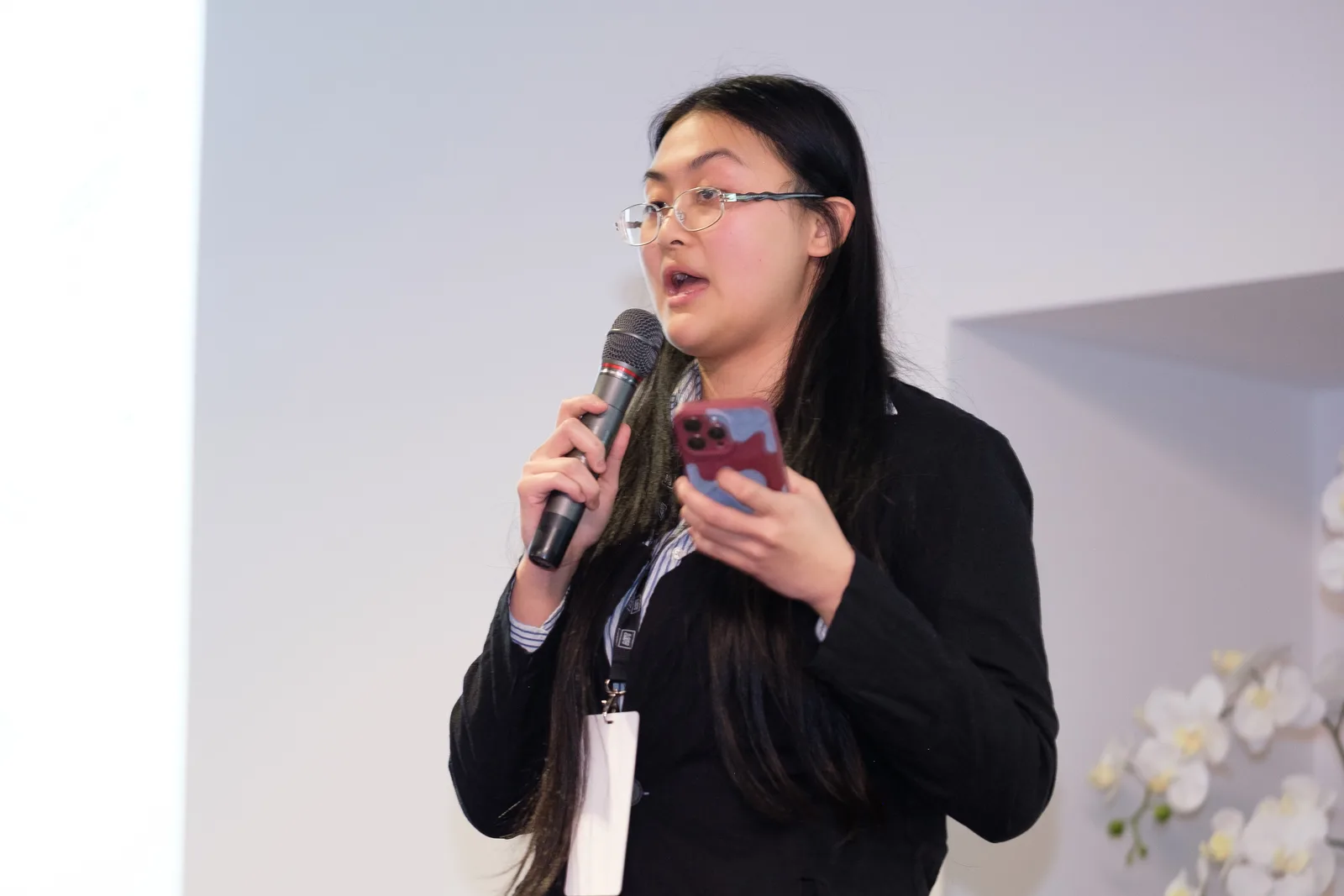 A young woman with long dark hair speaks into a handheld microphone against a plain white wall, her phone held in her other hand as she presents.