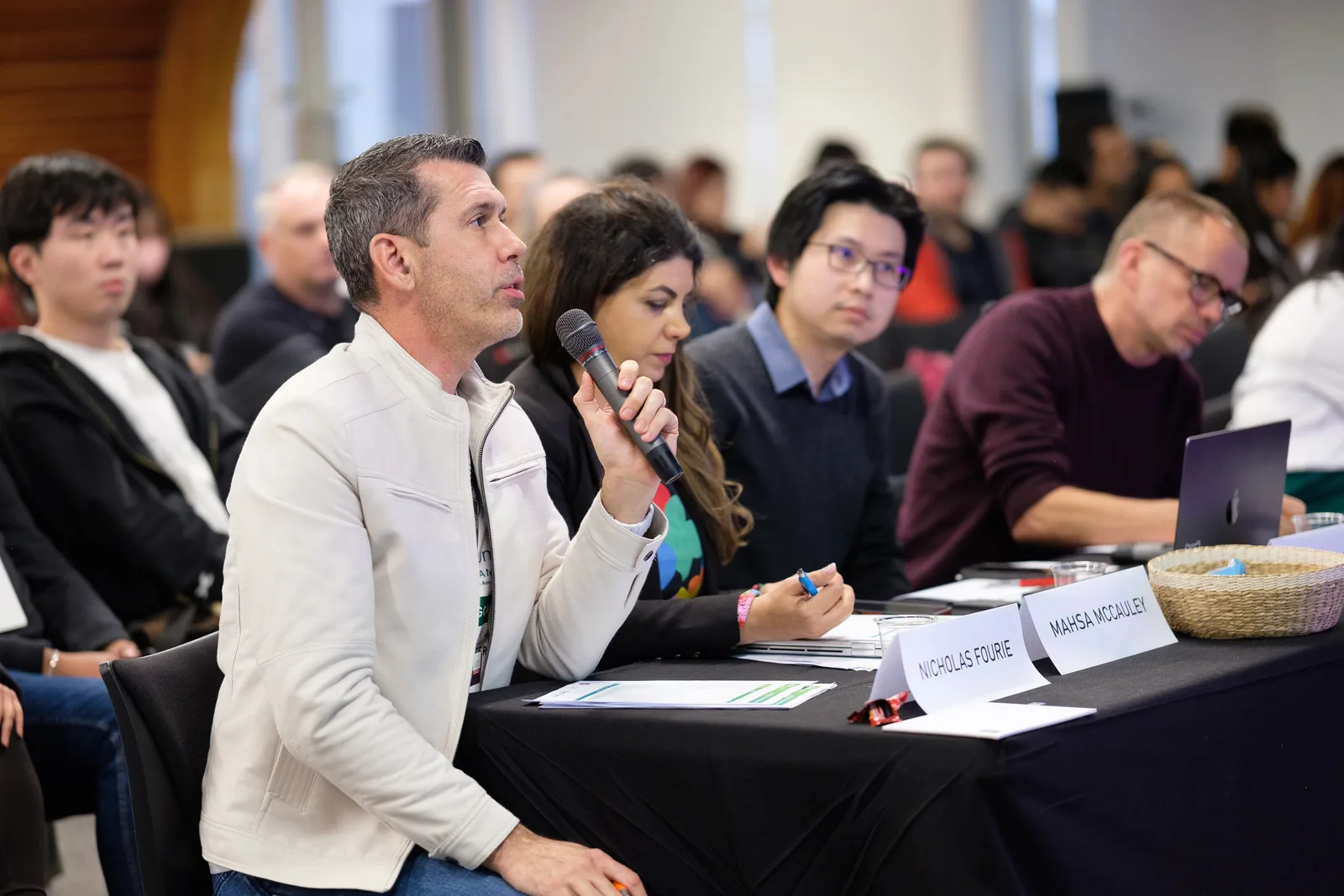 A judge lifts the microphone to question a team from the long panel table while the rest of the judges take notes and the audience watches behind.