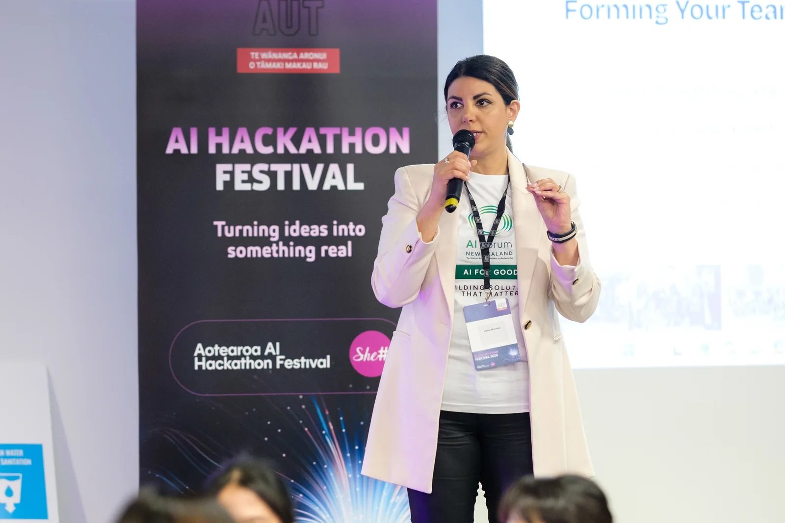 A speaker with a microphone in front of the Aotearoa AI Hackathon Festival banner reading 'Turning ideas into something real'