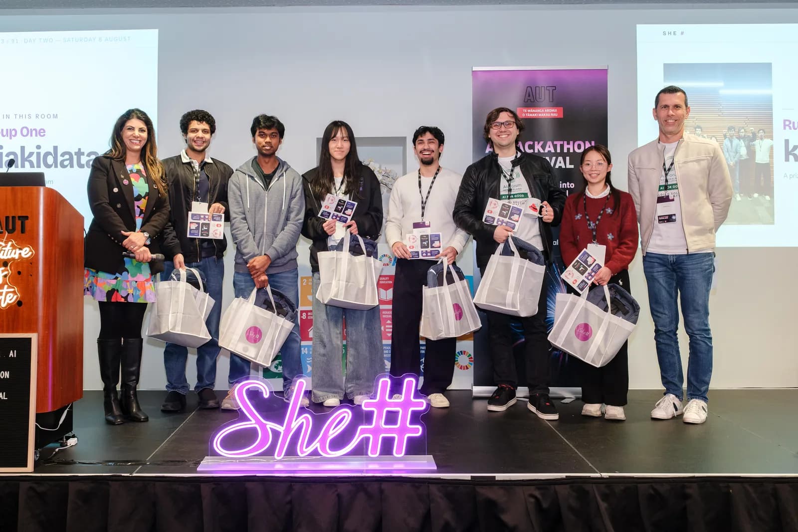A winning team on stage with their prize totes, behind the neon She Sharp sign