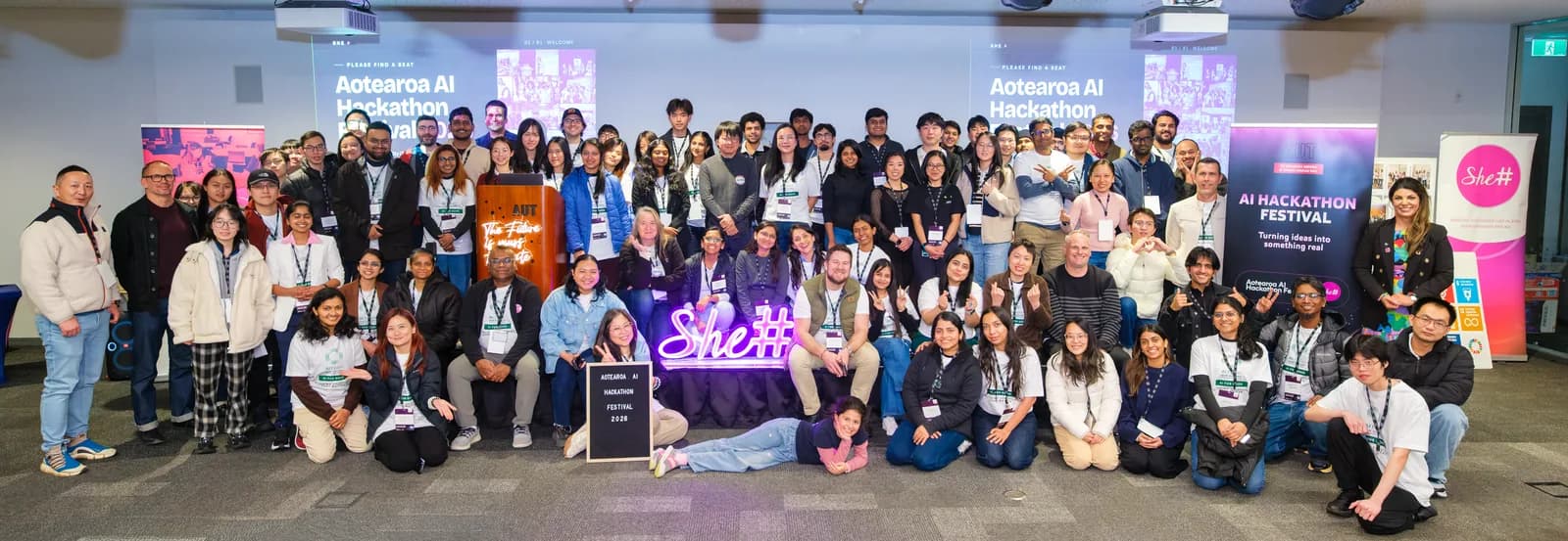 Around ninety participants and organisers massed together for a whole-festival group photograph, gathered around a neon She Sharp sign beneath the Aotearoa AI Hackathon Festival banners.