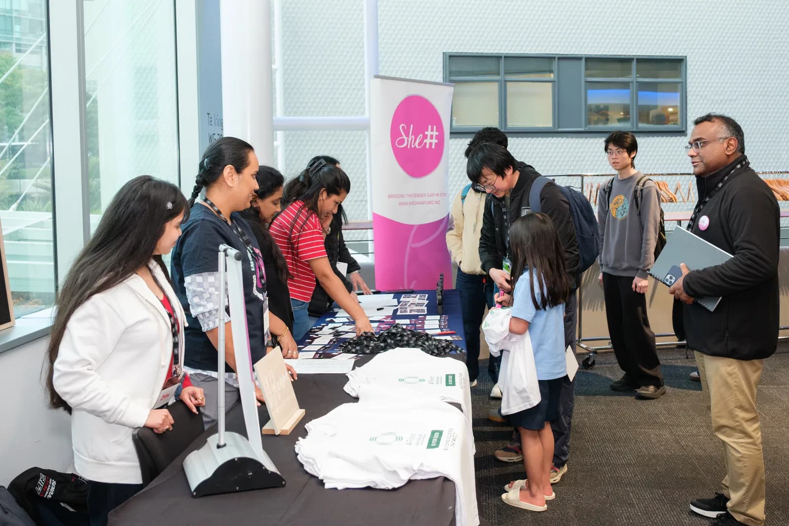 Attendees collect lanyards and folded white t-shirts from a welcome table beside a pink She# pull-up banner in a glass-walled campus foyer.