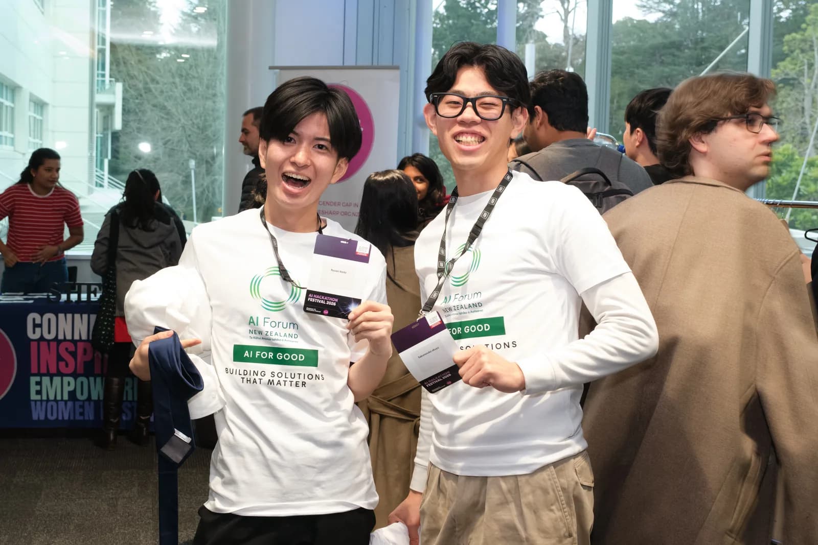 Two participants in white AI Forum New Zealand t-shirts reading AI FOR GOOD laugh together, holding up their festival name badges.