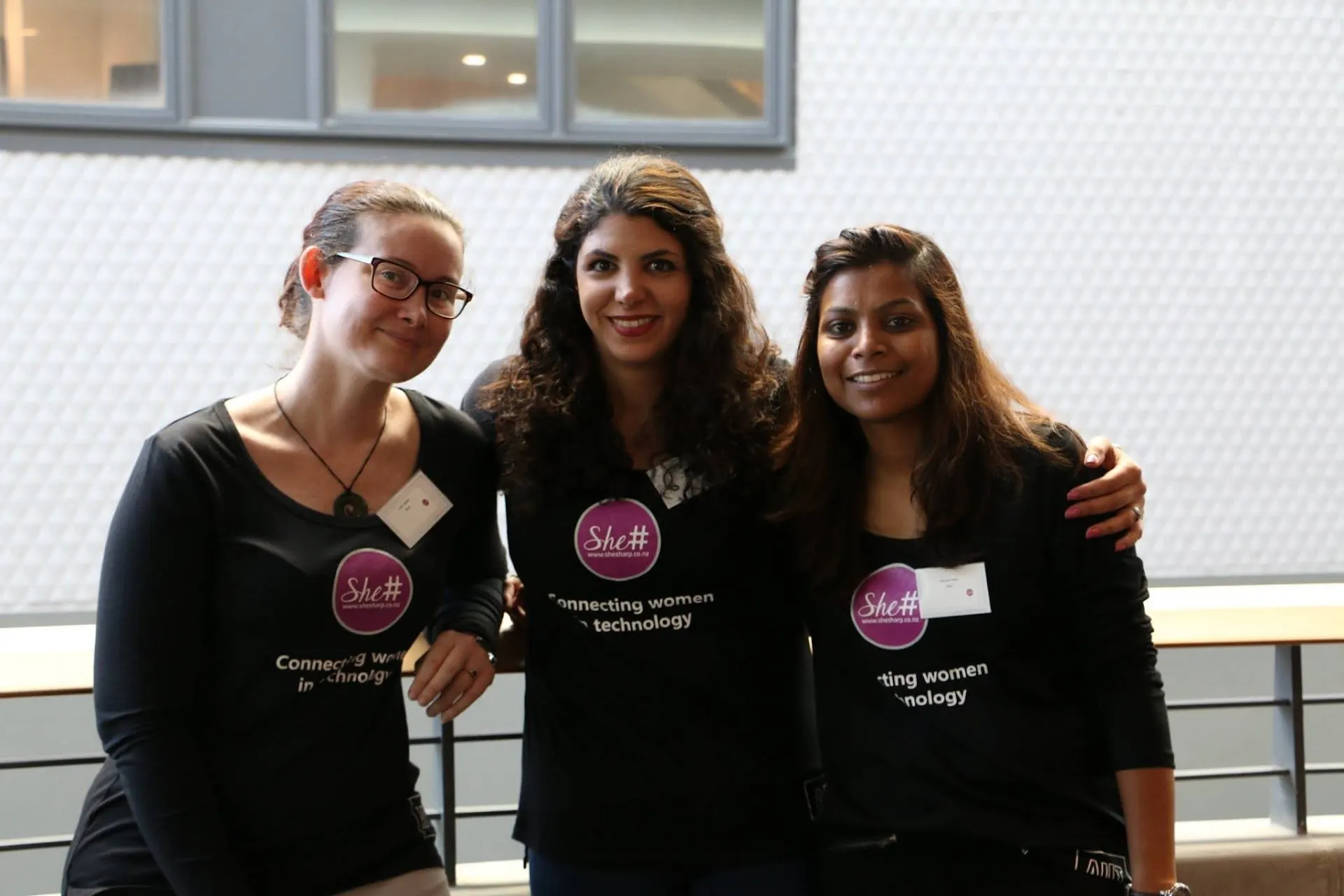 Three She Sharp volunteers in branded T-shirts standing arm in arm and smiling together at a Google event.