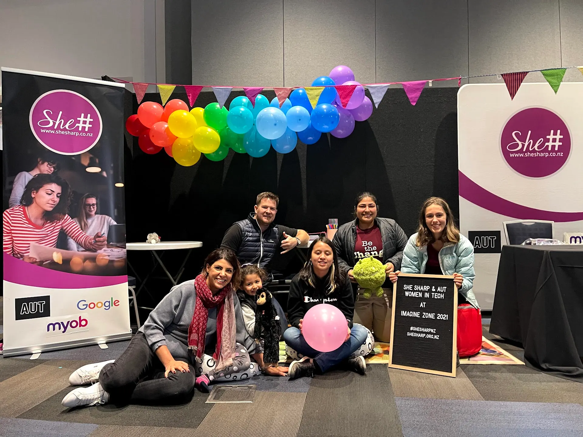 A group gathered on the floor in front of a stage decorated with rainbow balloons and bunting at the She Sharp Imagine Zone event.